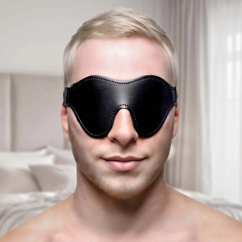 A person wears a black Strict Fleece Lined Blindfold, featuring a contoured leather-look design.