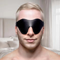 A person wears a black Strict Fleece Lined Blindfold, featuring a contoured leather-look design.