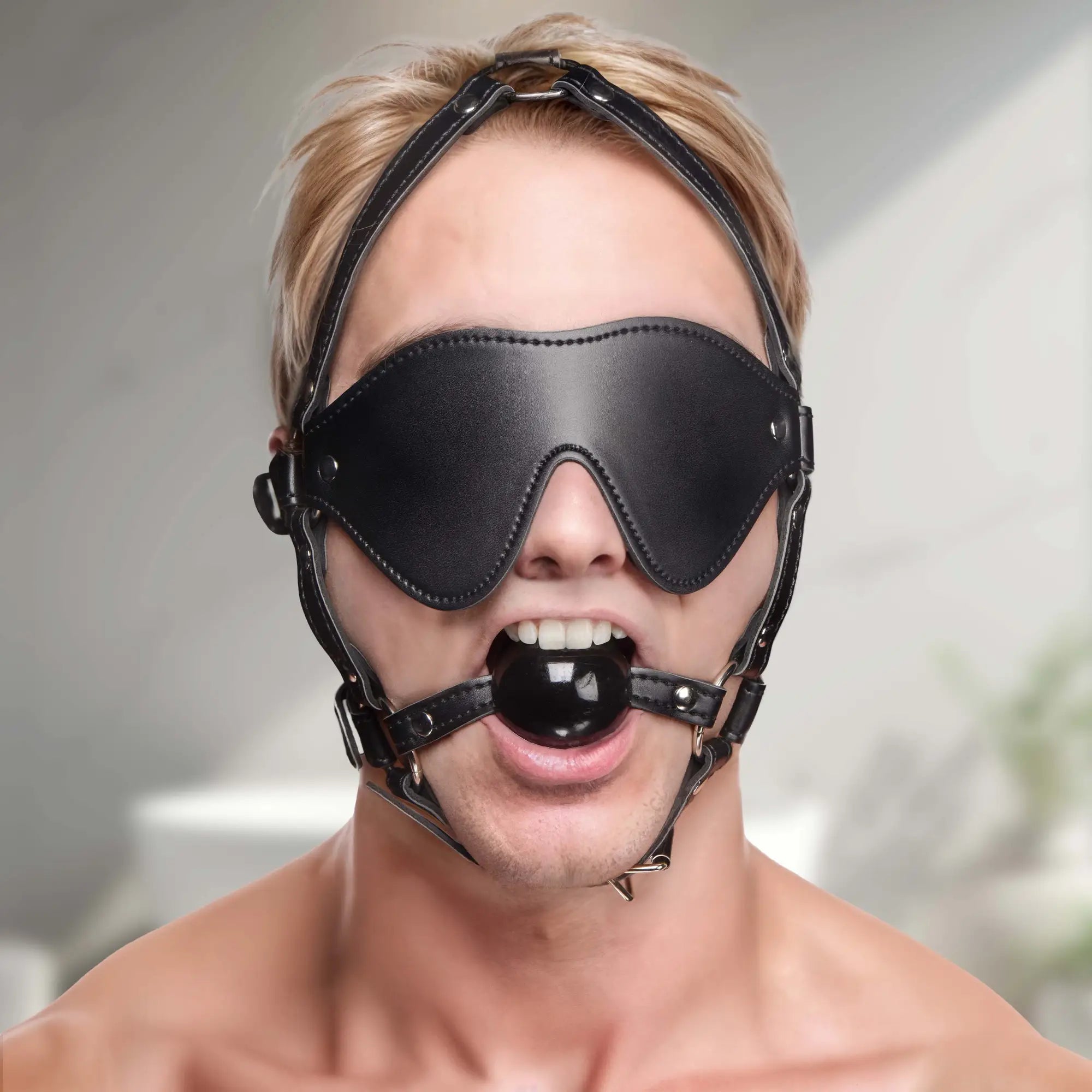 Strict Blindfold Harness and Ball Gag: A person wears a black leather blindfold, adjustable head harness, and ball gag.