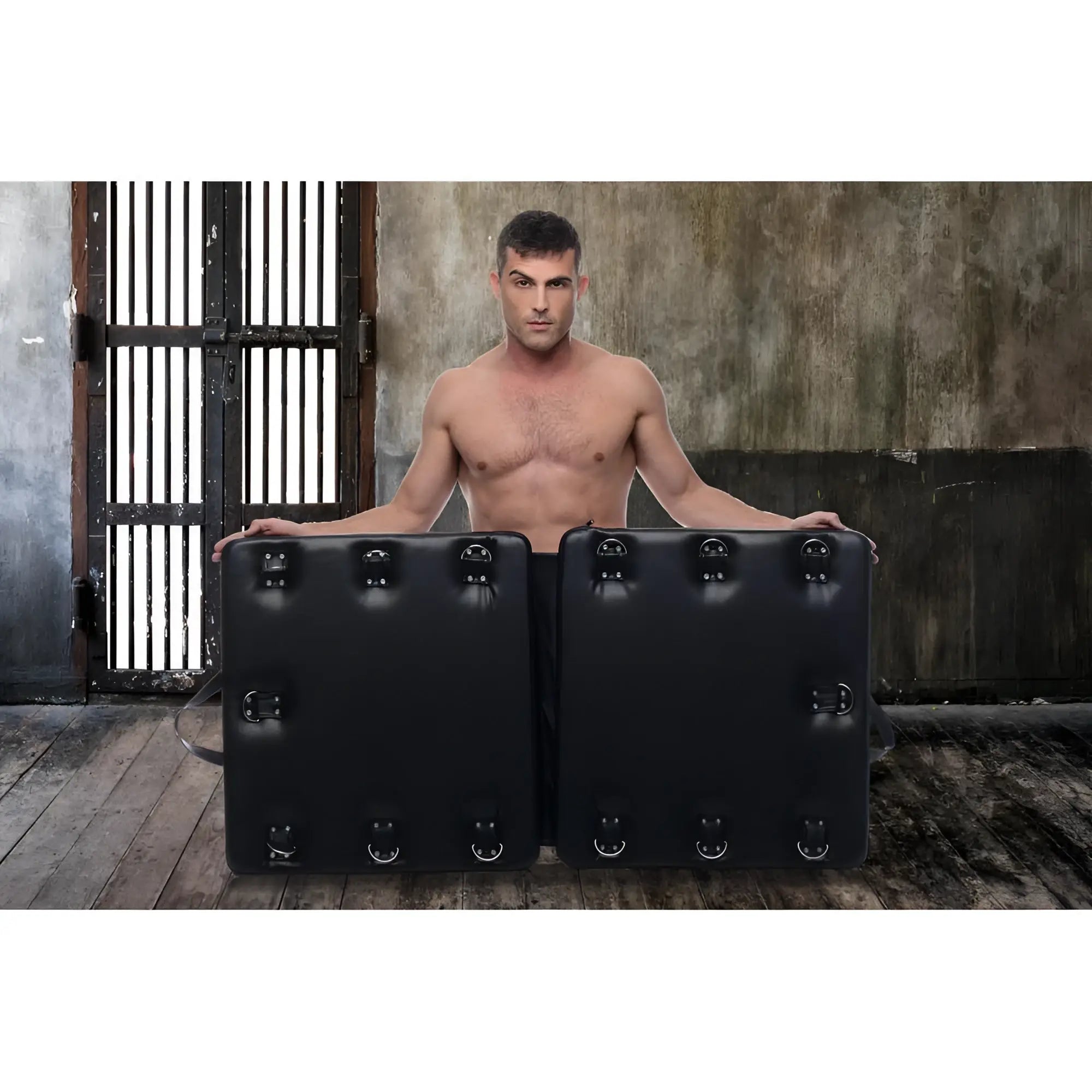 A muscular man stands behind a black, folding Strict Bondage Board featuring multiple metal D-ring attachment points.