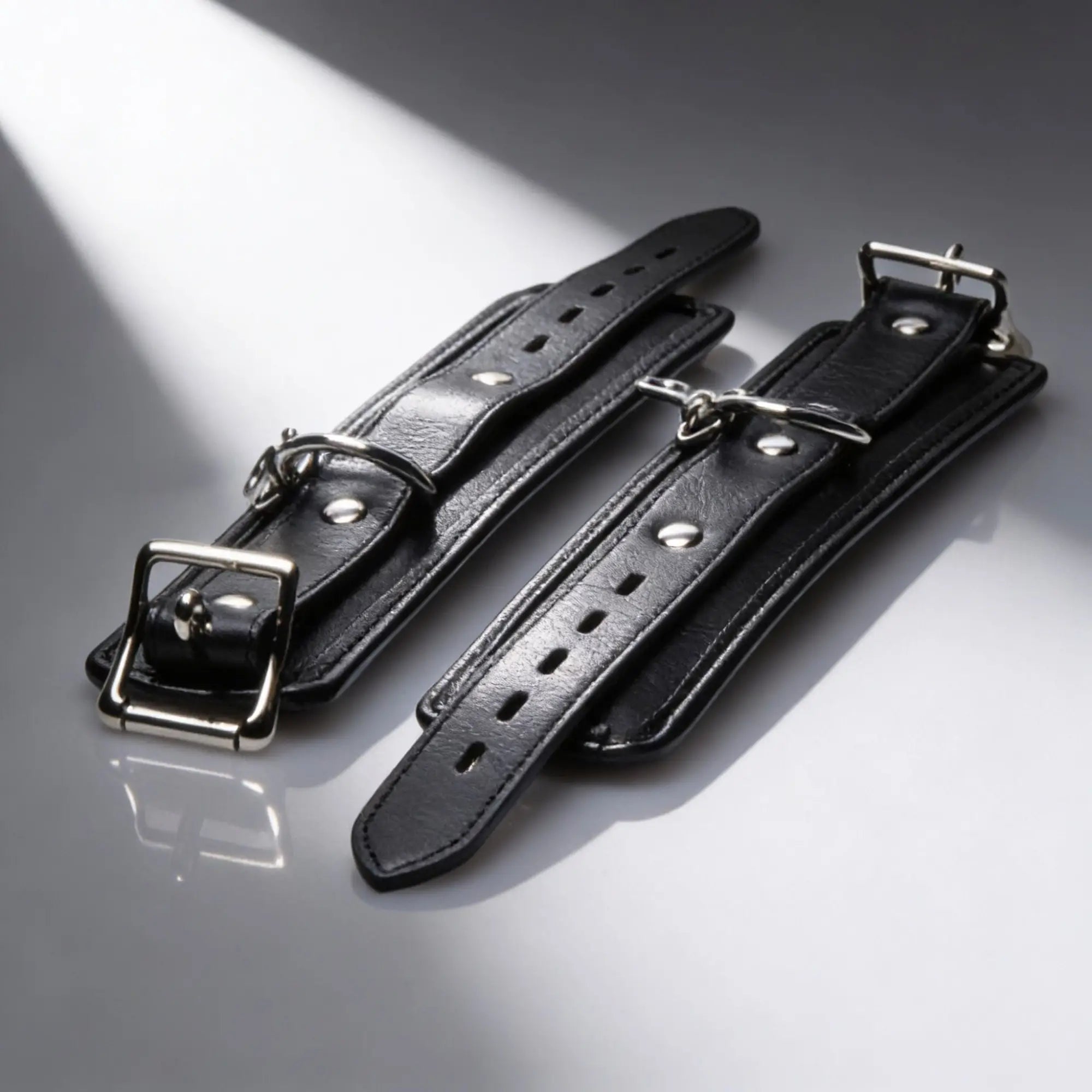 Strict Deluxe Bed Restraint System: Two black leather cuffs featuring silver buckles and D-rings on a white background.