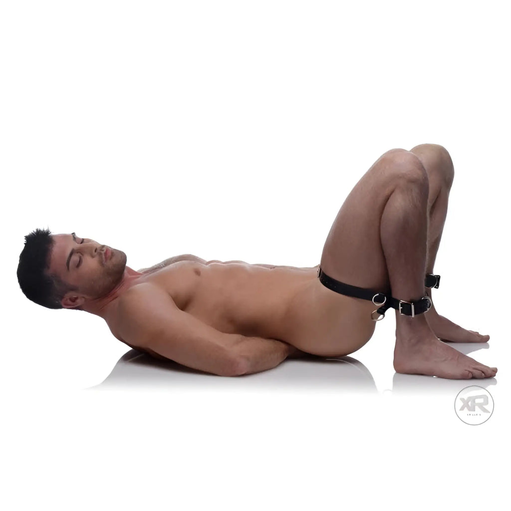 Strict Frog Tie Kneeling Bondage System: A man lying on his back with ankles cuffed to a black leather thigh strap.