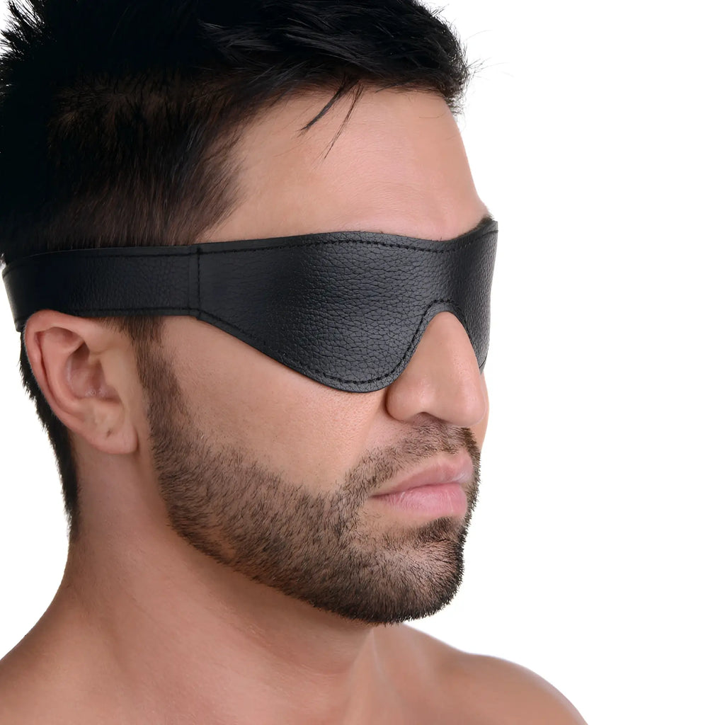 Strict Leather black leather blindfold worn by a man with dark hair and stubble, shown in profile.