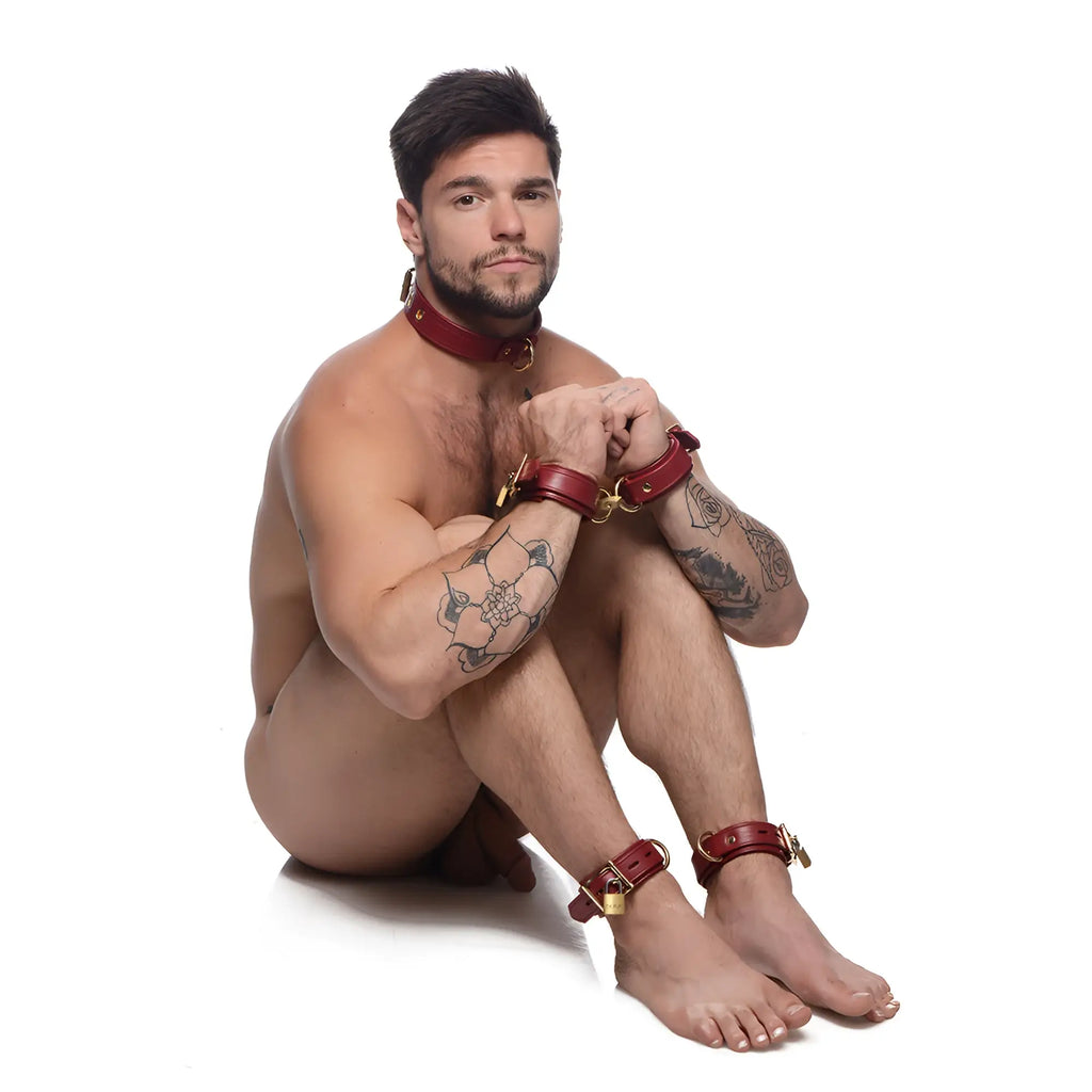 Strict Leather burgundy premium leather collar, wrist cuffs, and ankle cuffs with gold hardware on a tattooed man.