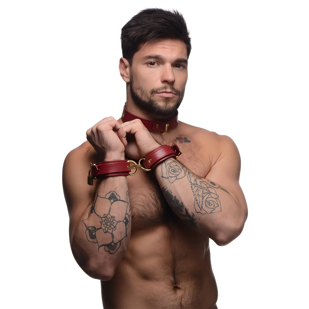 A shirtless man with tattoos wears a burgundy Strict Leather premium leather collar and locking wrist cuffs.