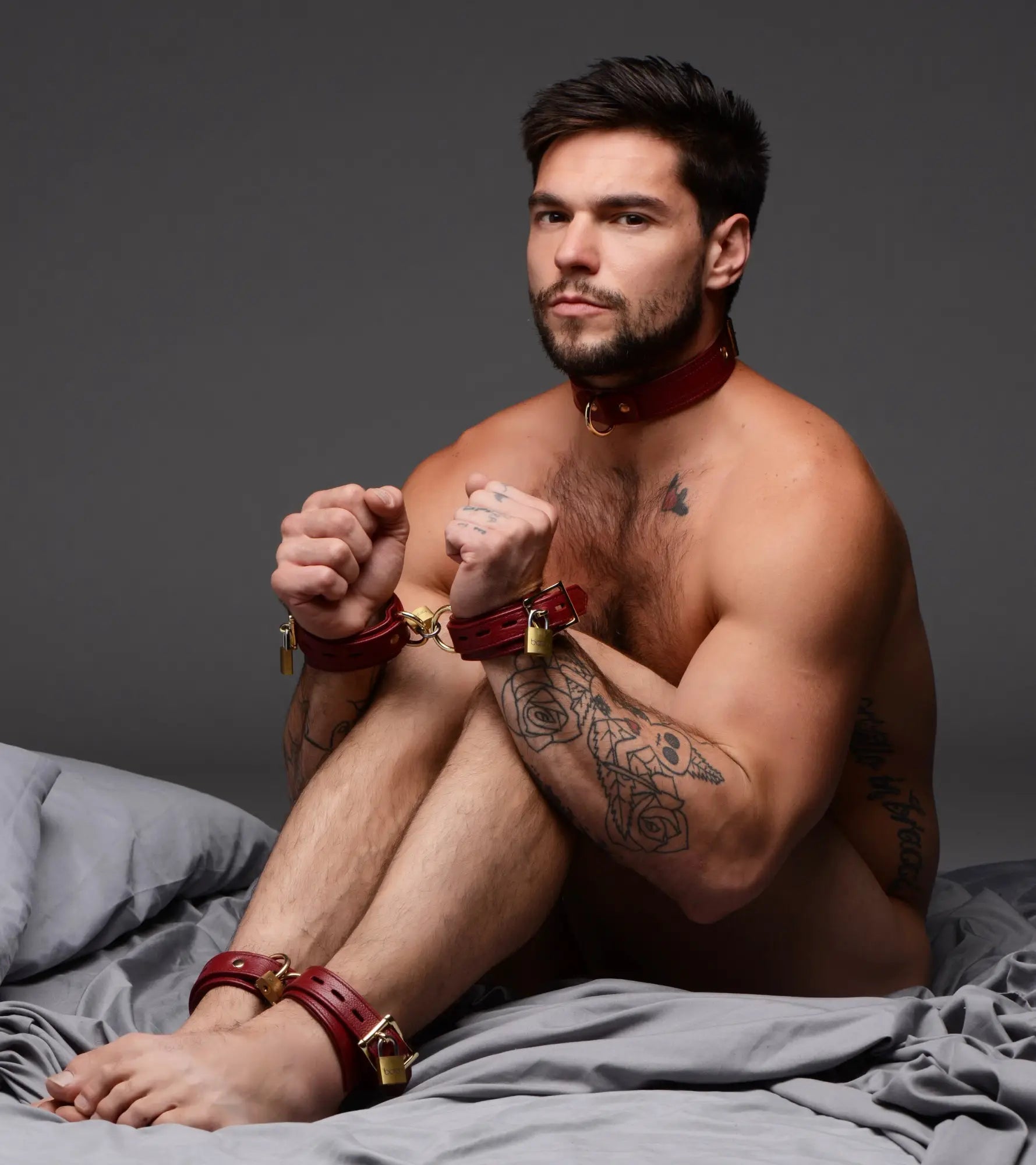 Strict Leather burgundy premium leather collar, wrist, and ankle cuffs with gold hardware and locks on a seated man.