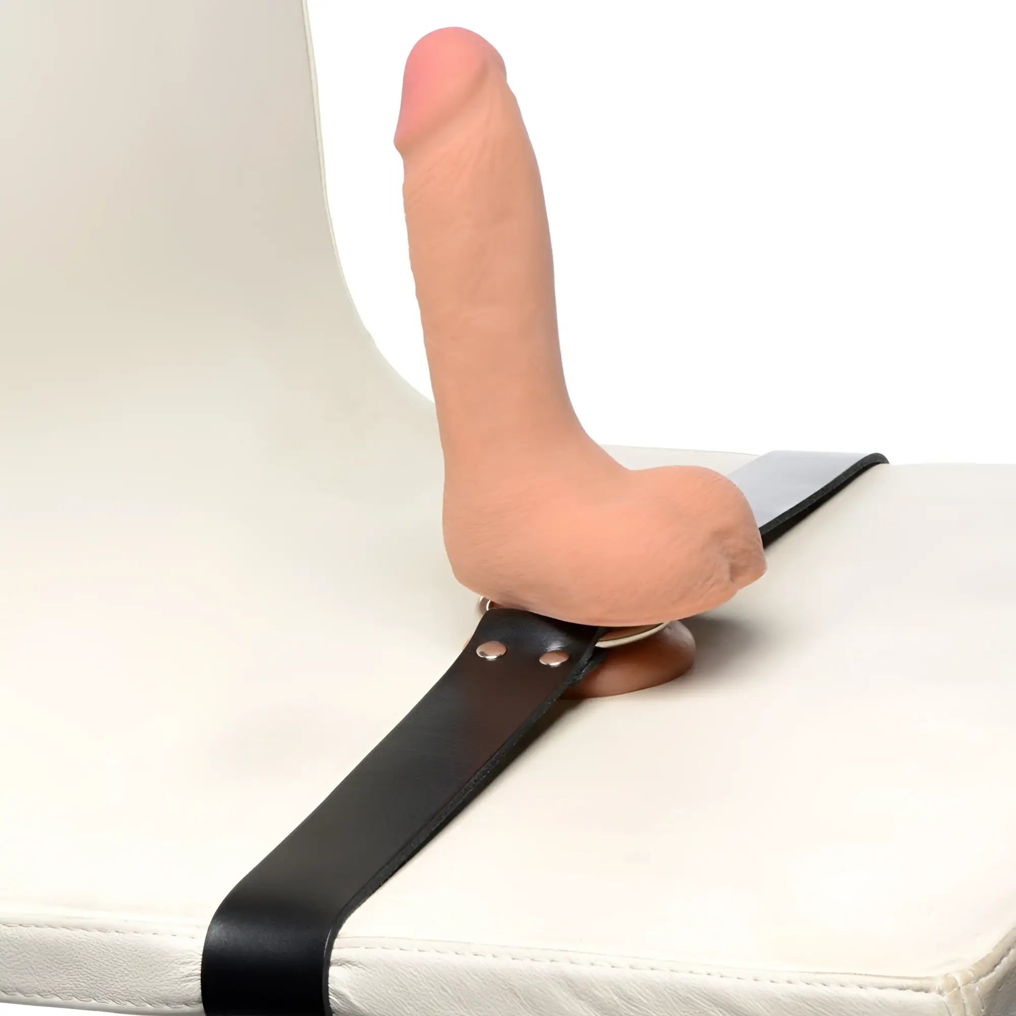 Strict Leather dildo strap featuring a realistic skin-toned dildo attached to a black leather belt on a white chair.