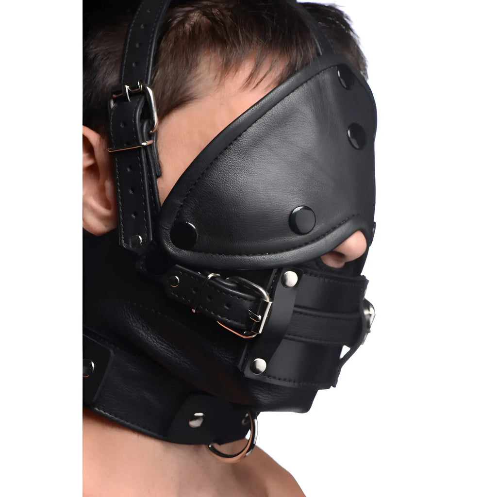 Strict Leather black leather head harness with buckle straps, eye covering, and a removable gag worn by a person.