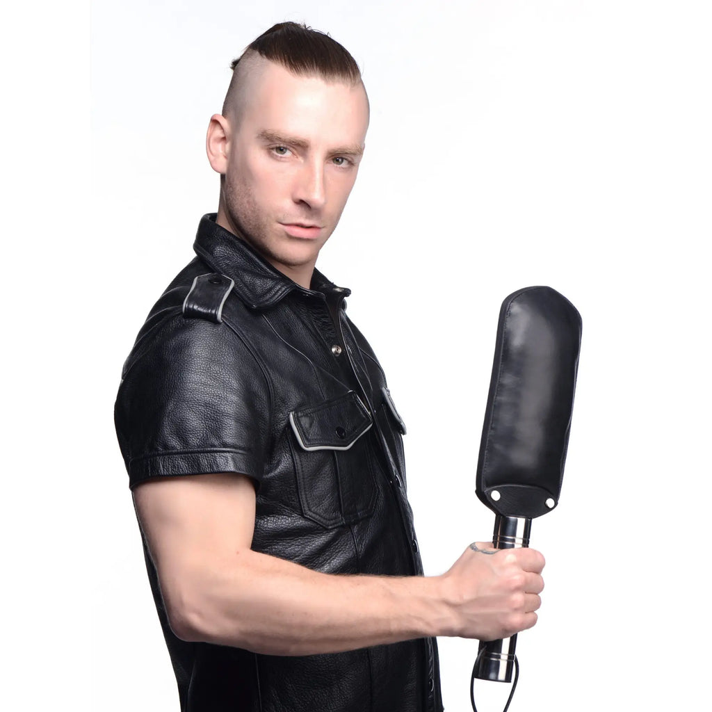 A man in a black leather shirt holding a Strict Leather padded paddle with a black leather head and metallic handle.