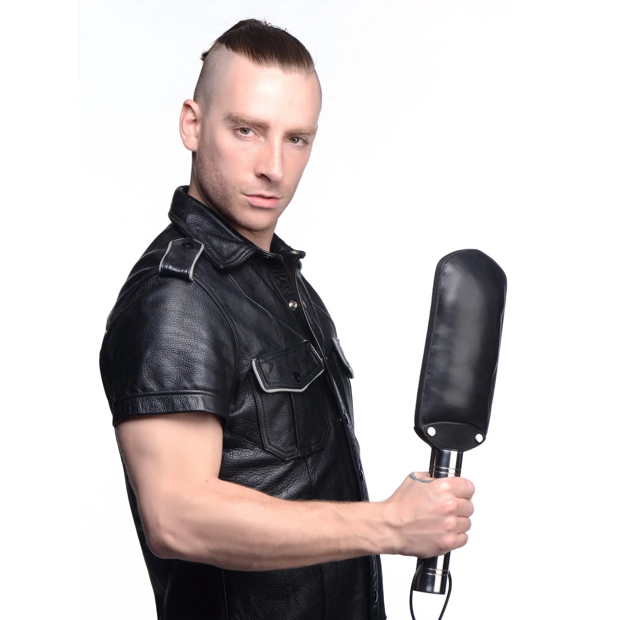 A man in a black leather shirt holding a Strict Leather padded paddle with a black leather head and metallic handle.