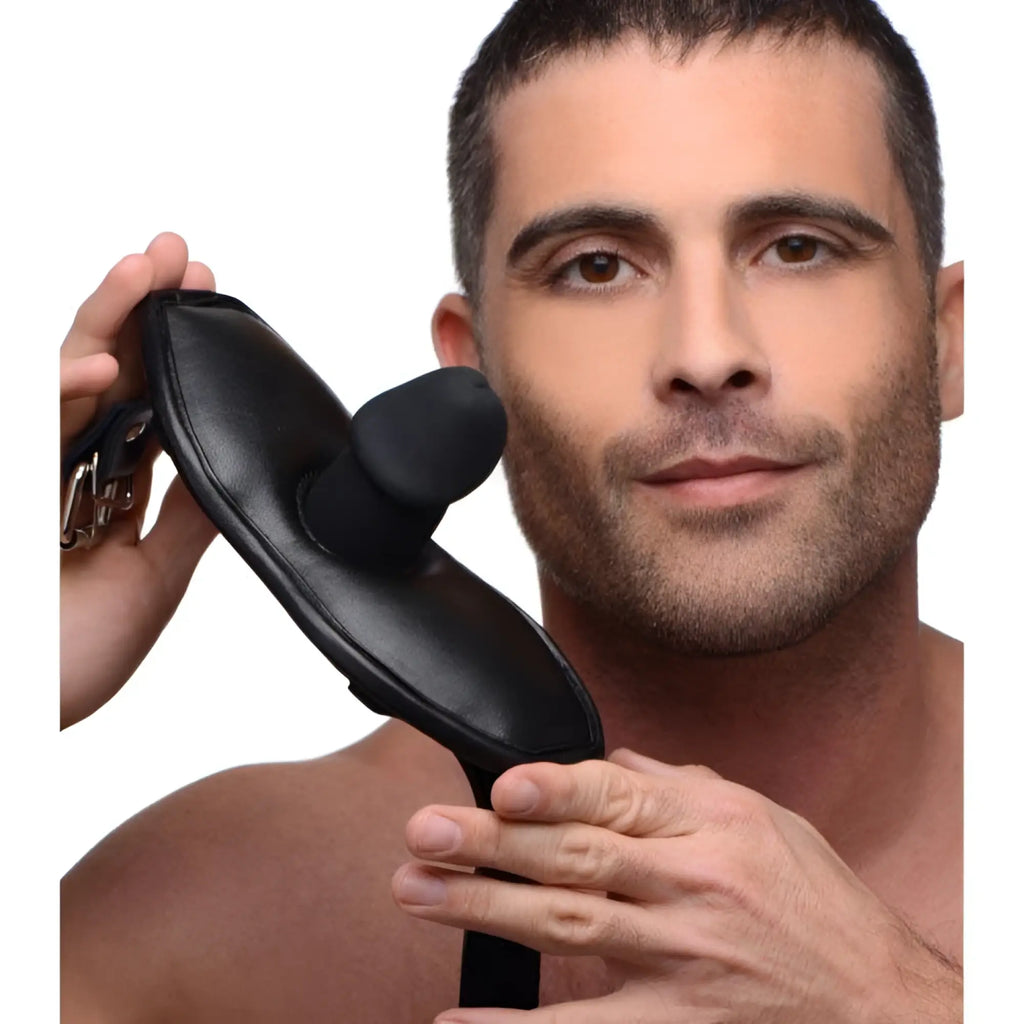 A man holds a Strict Leather black, padded silicone penis mouth gag with adjustable straps against a white background.