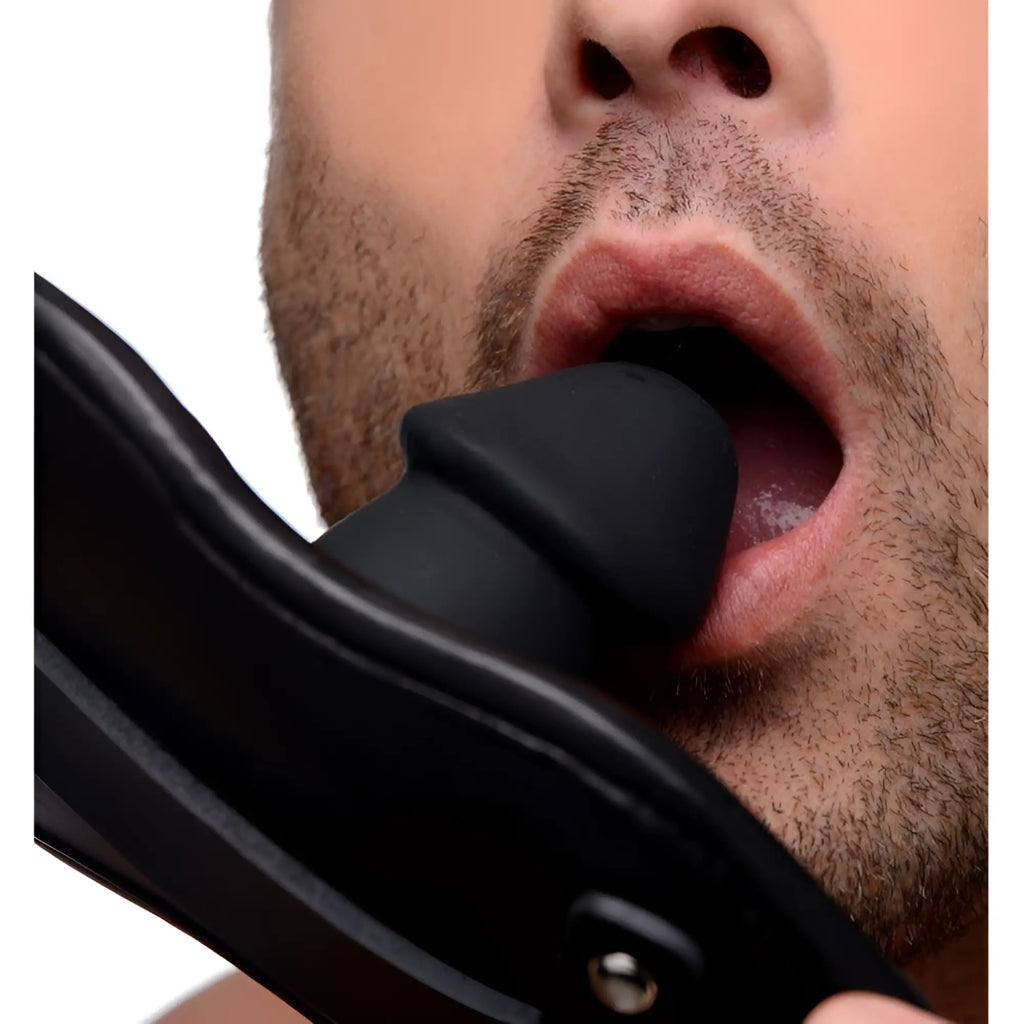 Strict Leather penis-shaped silicone mouth gag with a black padded leather strap, shown in use against a man’s mouth.