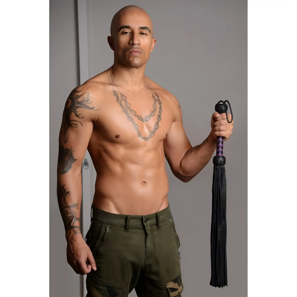 A shirtless, tattooed man holds a black Strict Leather Premium Deerskin Flogger with purple handle accents.