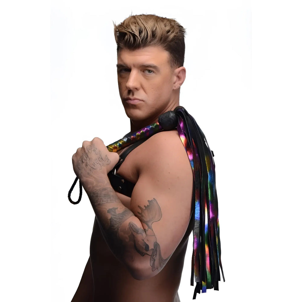 Strict Leather Rainbow Flogger with iridescent rainbow and black leather tails, held by a shirtless tattooed man.