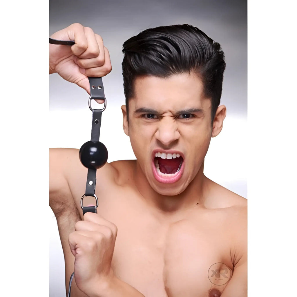 Strict Leather silicone ball gag with adjustable black leather straps, held by a shirtless man with an open mouth.