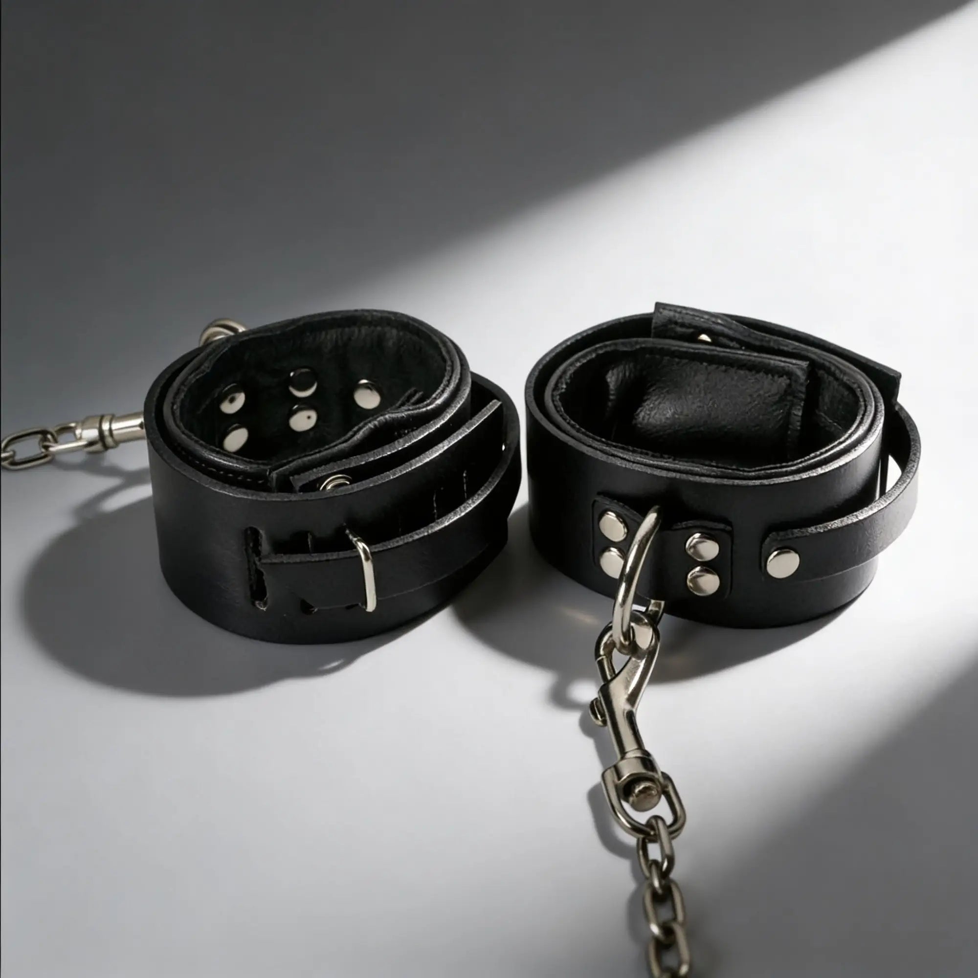 Strict Leather sling and stirrups: Two black leather ankle cuffs with silver-tone metal hardware and connecting chains.