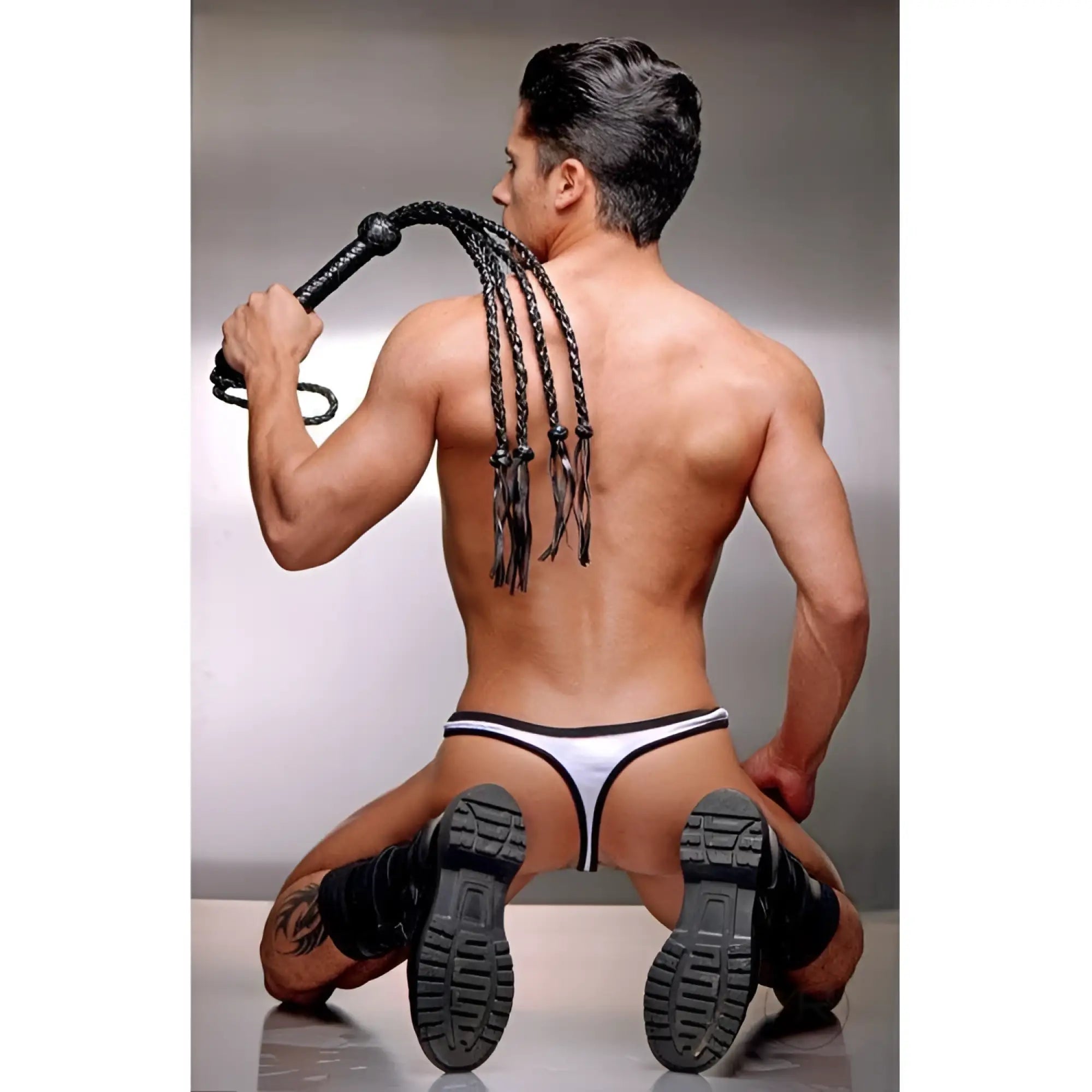 A man in a white thong and black boots holds a black Strict Leather Tigress whip with braided tails over his shoulder.