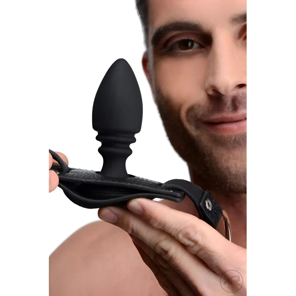 A man holds a black Strict silicone anal plug attached to a matching leather-textured cock ring harness.