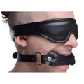 Strict Padded Blindfold and Gag Set: Black leather-style, padded blindfold and matching mouth gag on a person's face.