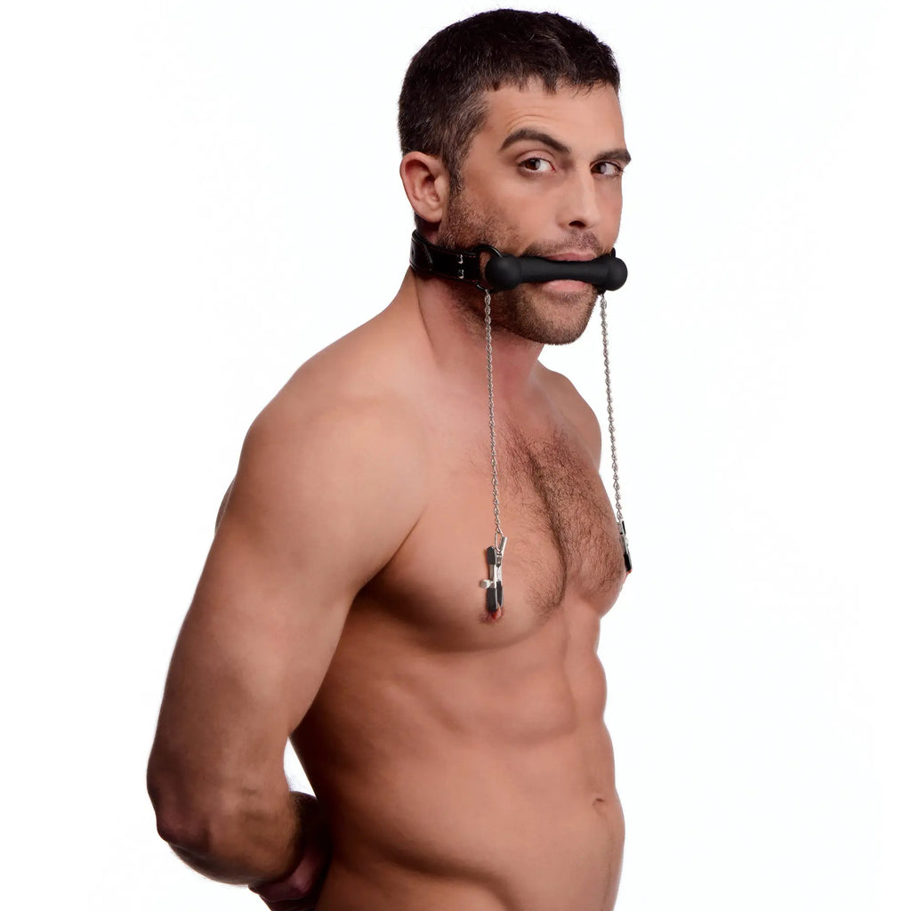 A man wearing a black Strict silicone bit gag with silver chains attached to metal nipple clamps.