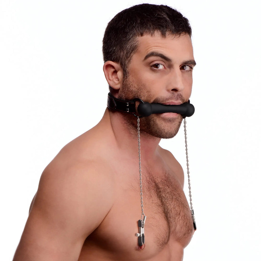 Man wearing a black Strict Silicone Bit Gag with silver chains connecting to black nipple clamps on a white background.