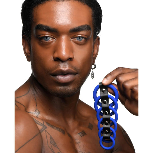 A man holds a Strict Silicone Gates of Hell chastity device, featuring blue rings linked by a black studded strap.
