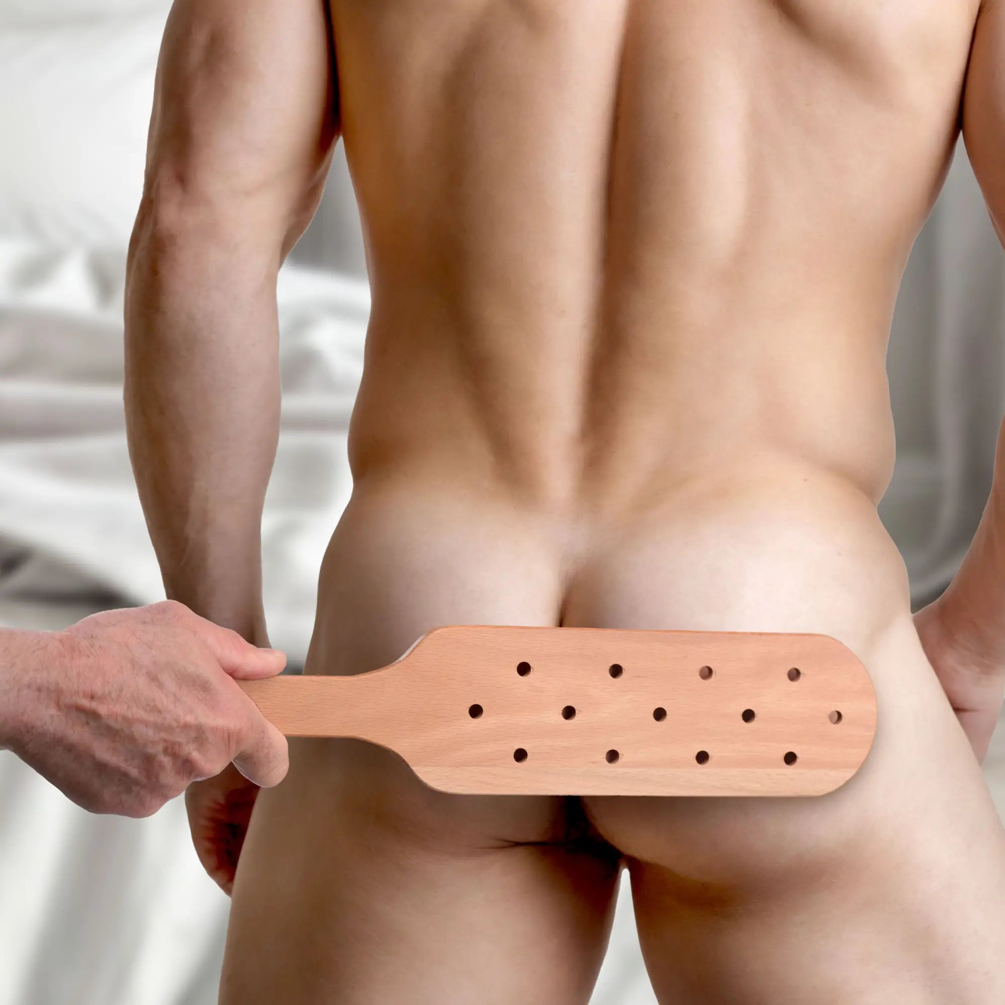 A hand holding a light brown Strict Wooden Paddle with perforated holes positioned against a person’s back.