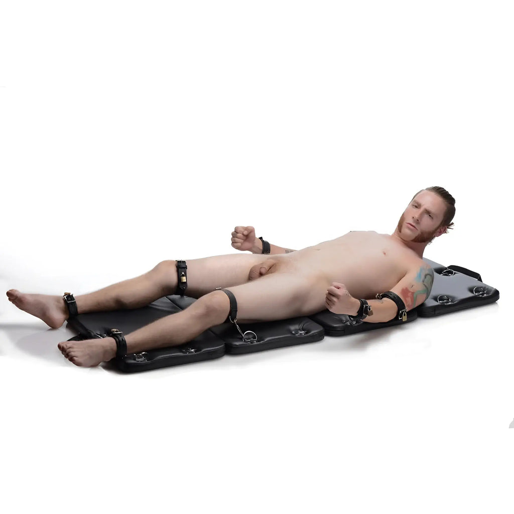 Strict XL Bondage Board: A man lies restrained on a black, padded mat with locking leather cuffs.