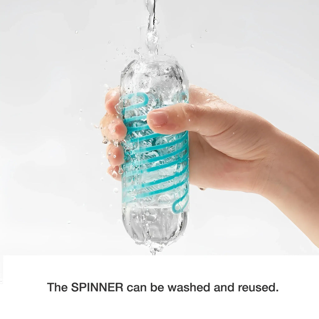 A hand rinses a clear Tenga Spinner 01 Tetra Stroker with a blue spiral internal texture under running water.
