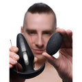 A man holds the black Thump It Kinetic Thumping 7X Large Anal Plug and its remote control.