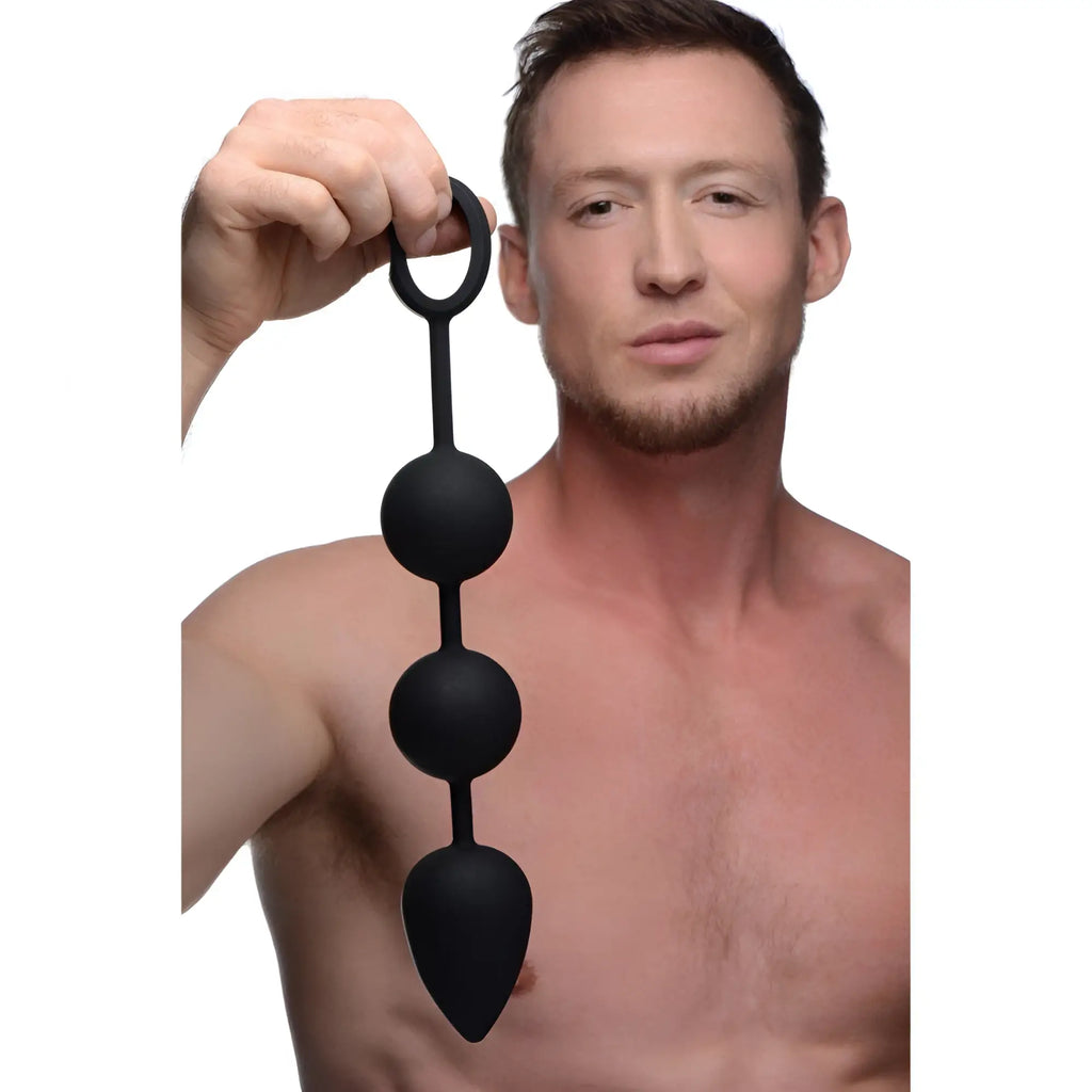 A shirtless man holds a black, weighted Tom of Finland silicone anal bead set featuring a circular pull ring.