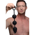 A shirtless man holds a black, weighted Tom of Finland silicone anal bead set featuring a circular pull ring.
