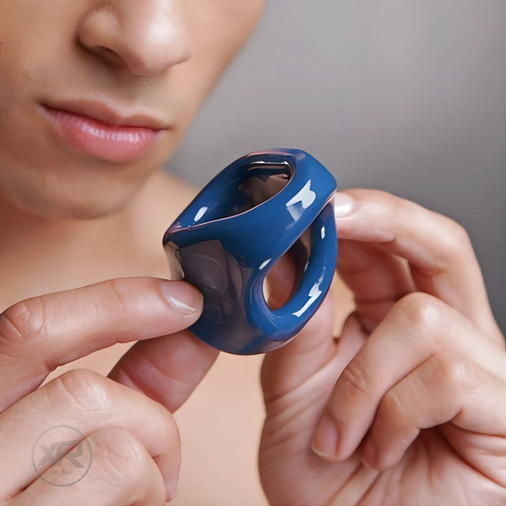 A person holds a glossy, dark blue Trinity dual cock and ball ring featuring a smooth, sculpted ergonomic design.