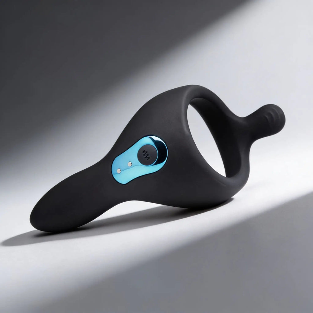 Trinity Men Power Taint 7X: Matte black silicone ring with a metallic blue control panel on a smooth, angled surface.