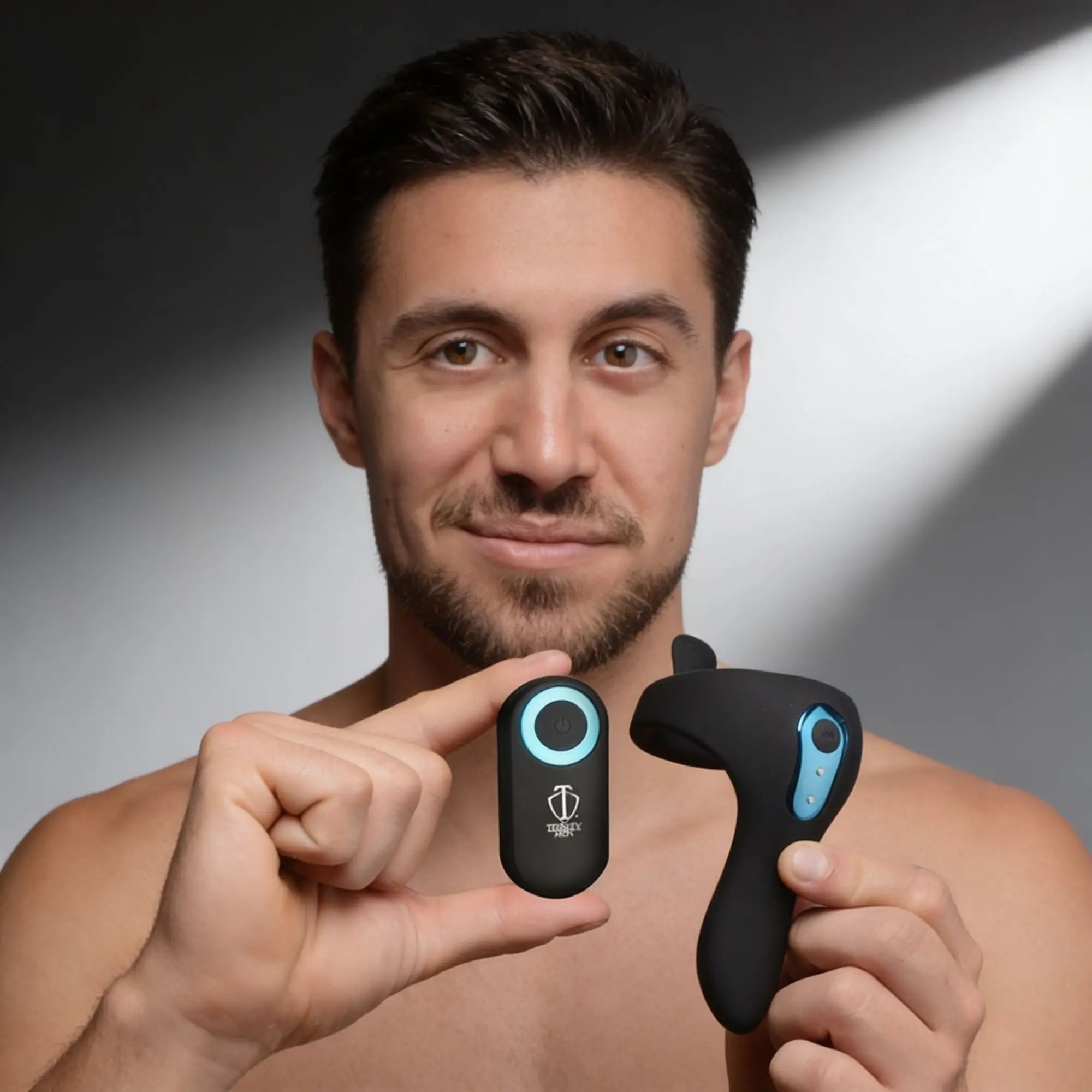 A man holding the black Trinity Men Power Taint 7X silicone cock ring and its matching remote with blue accents.