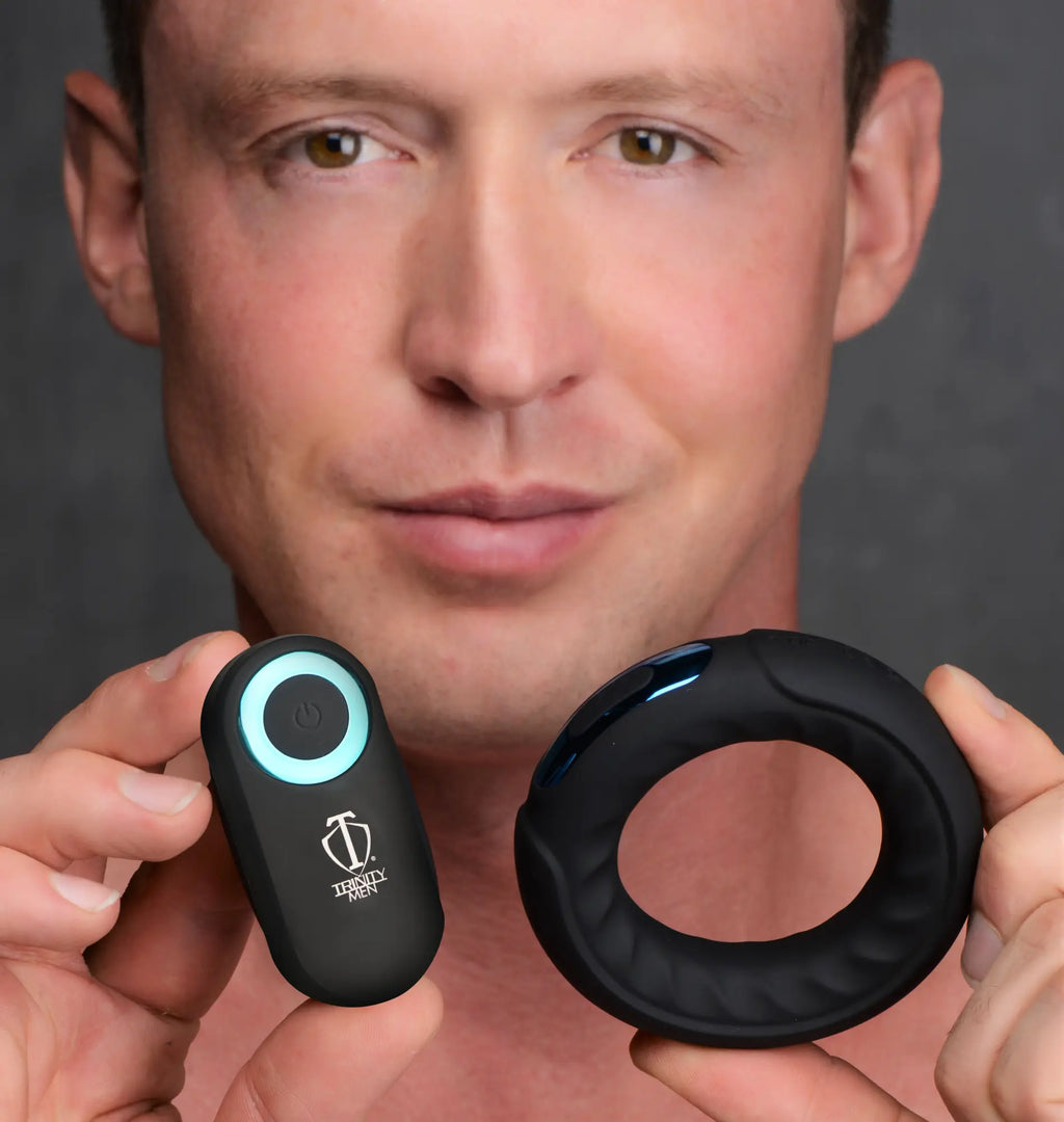 A man holds the black, silicone Trinity Men Remote Control 7X cock ring and its matching remote with a glowing LED.