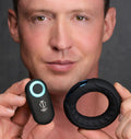 A man holds the black, silicone Trinity Men Remote Control 7X cock ring and its matching remote with a glowing LED.