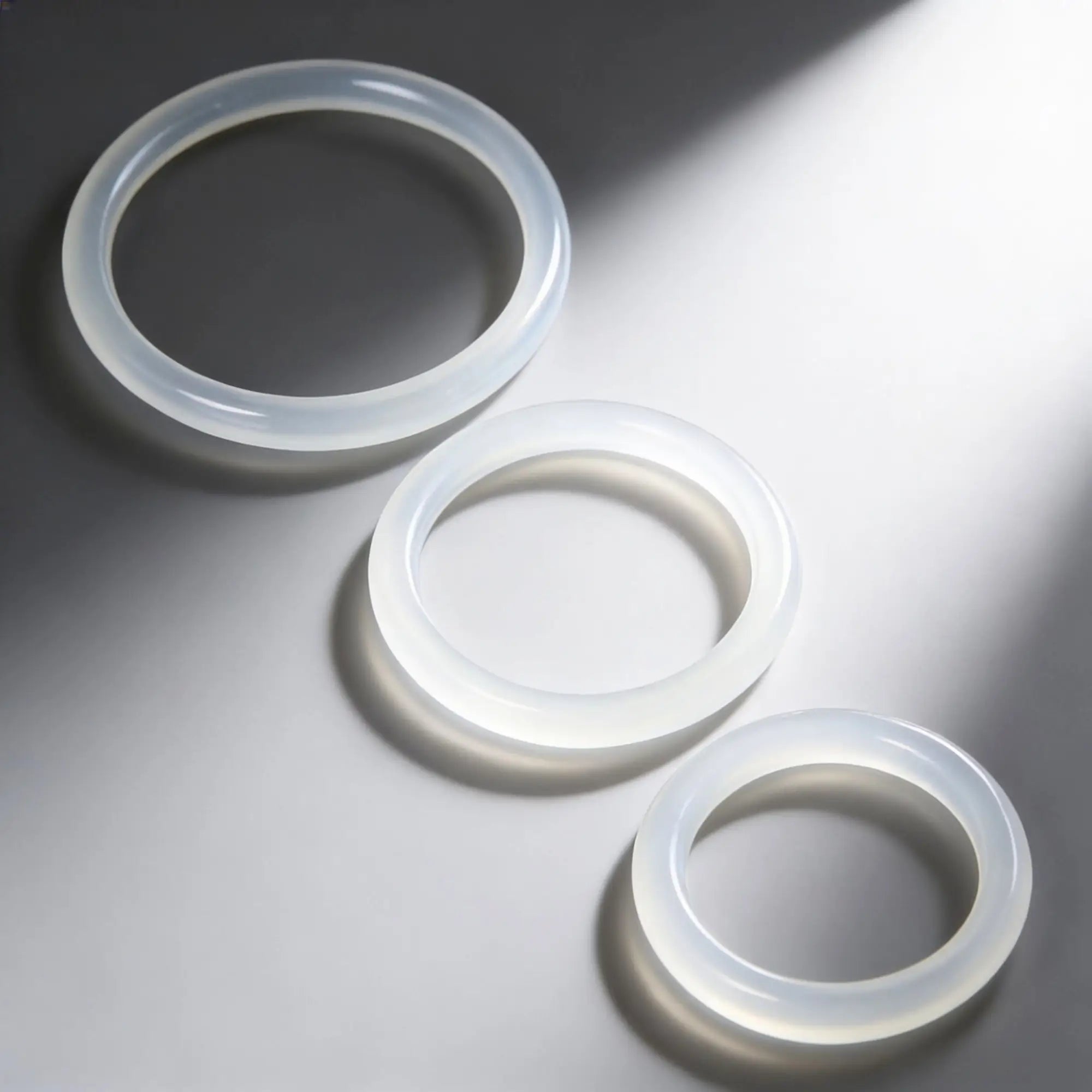 Trinity Men Silicone Cock Rings: Three smooth, translucent white rings of varying sizes arranged on a gray background.