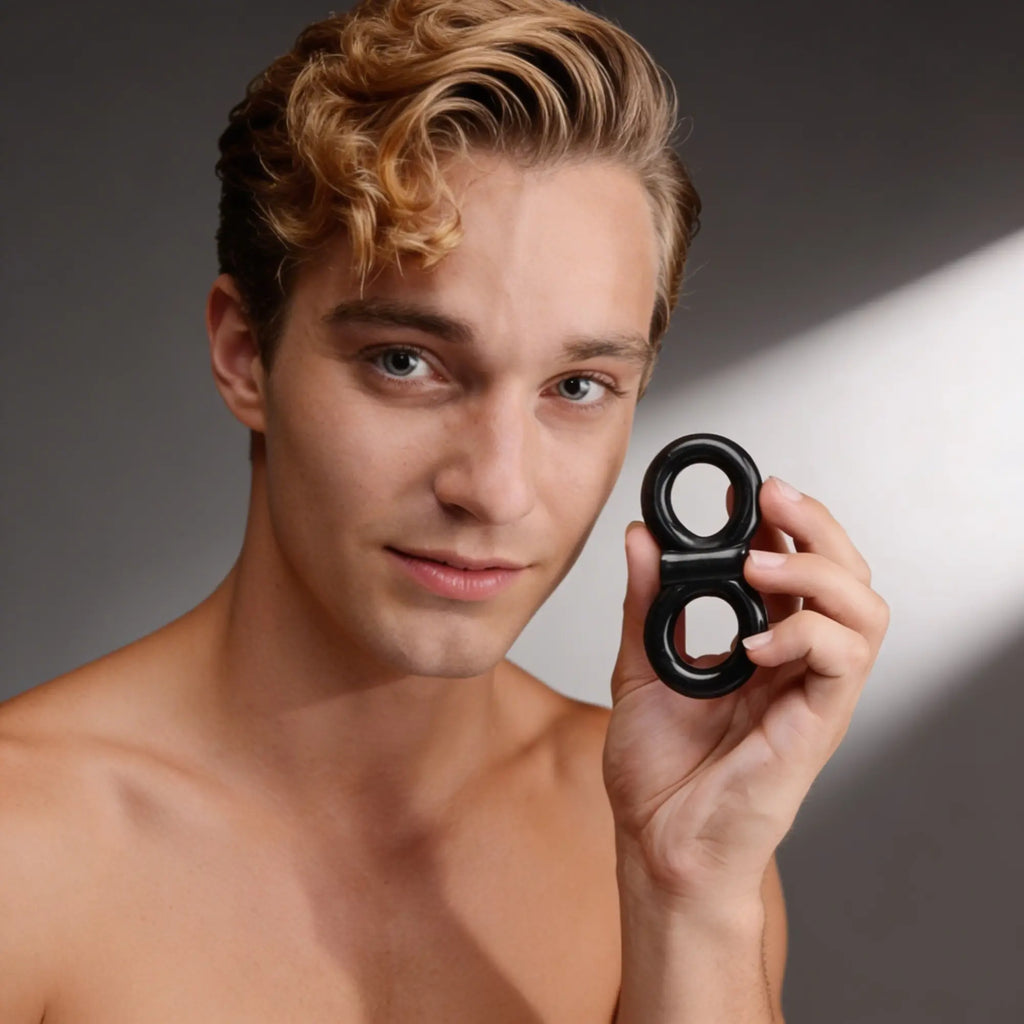 A man holds a smooth, black Trinity Men vibrating double cock and ball ring in his hand against a grey background.