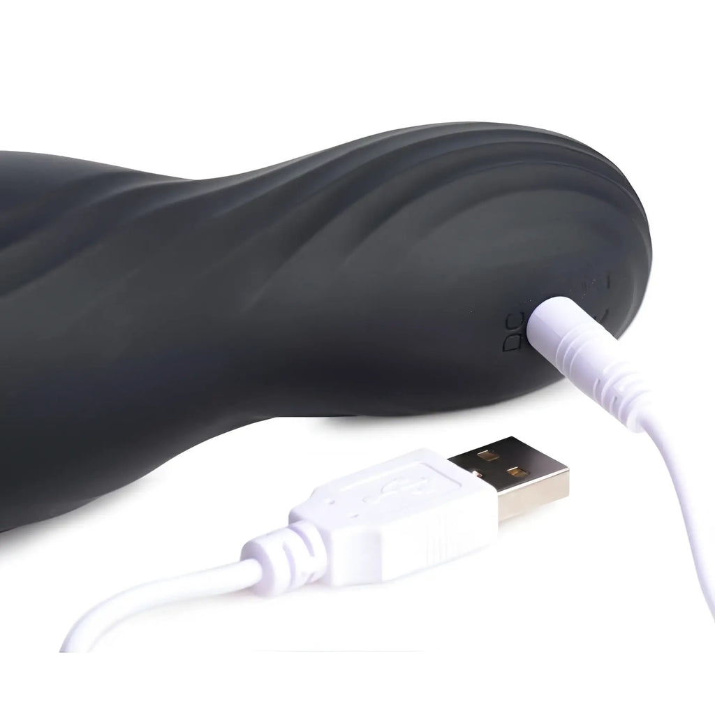Trinity Men Vibrating Rechargeable Penis Pleaser: A matte black, contoured device being charged via a white USB cable.