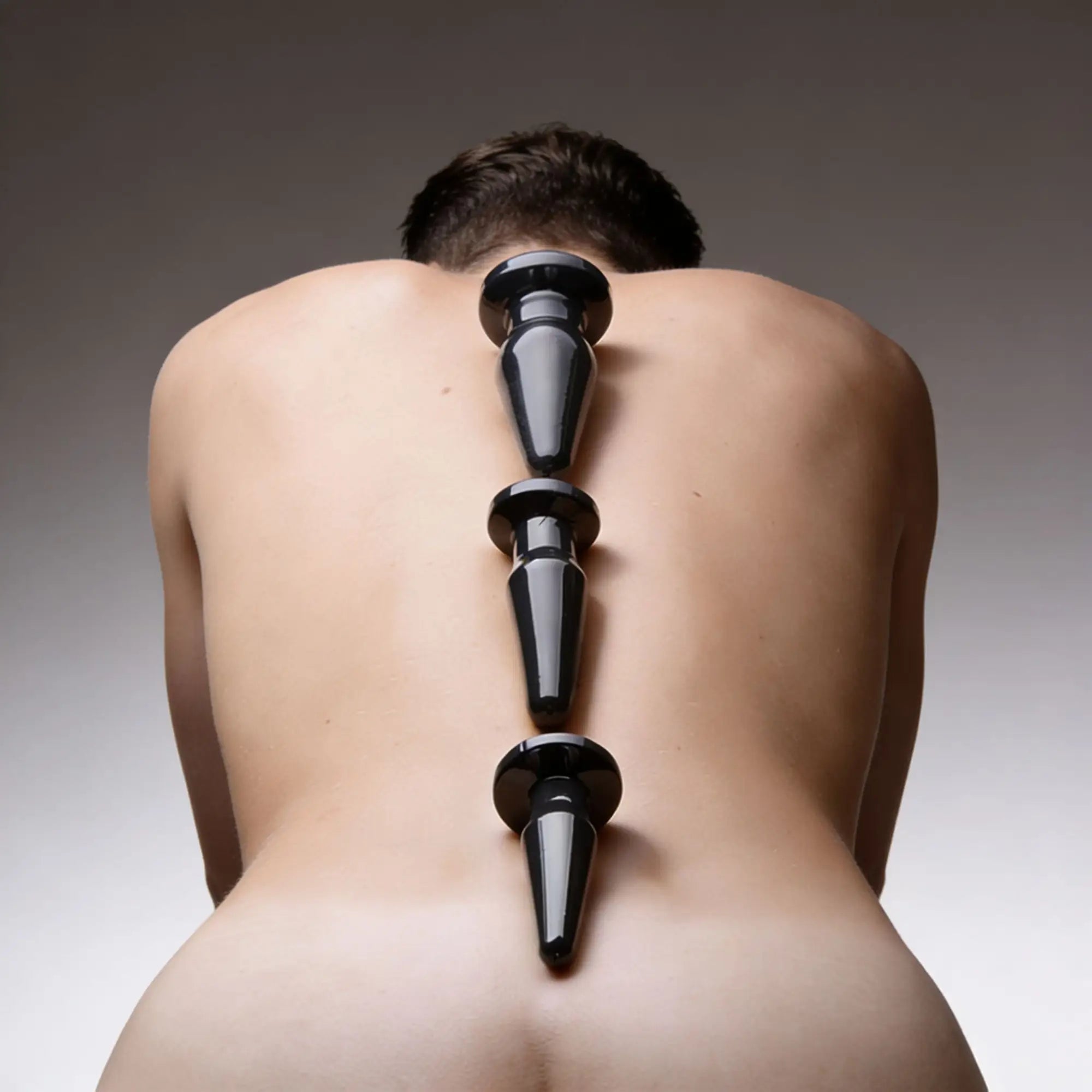 Trinity Vibes Anal Trainer Set featuring three glossy black, tapered plugs aligned down a person's bare back.