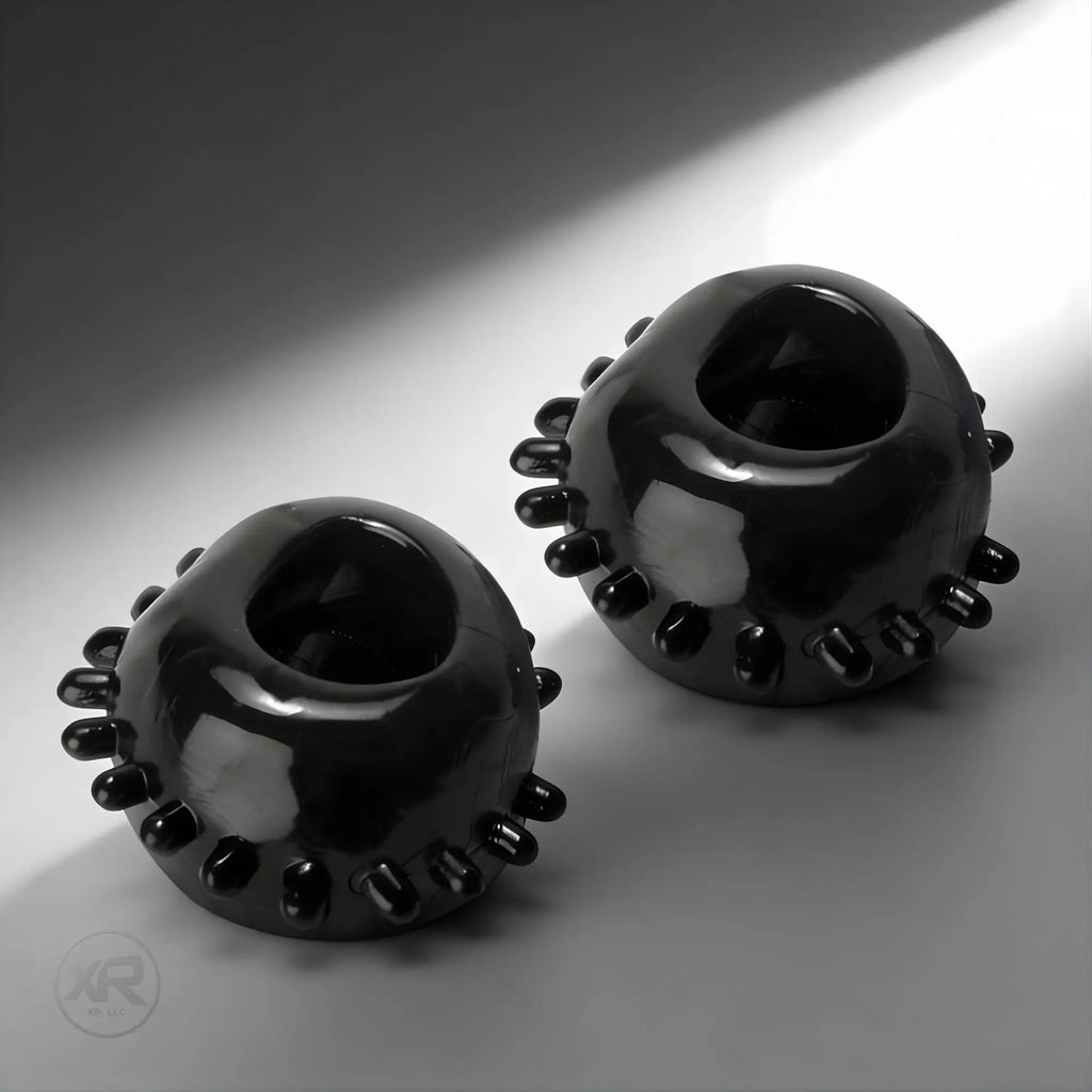Two shiny black Trinity Vibes gummy cock rings with small textured studs, displayed against a grey background.