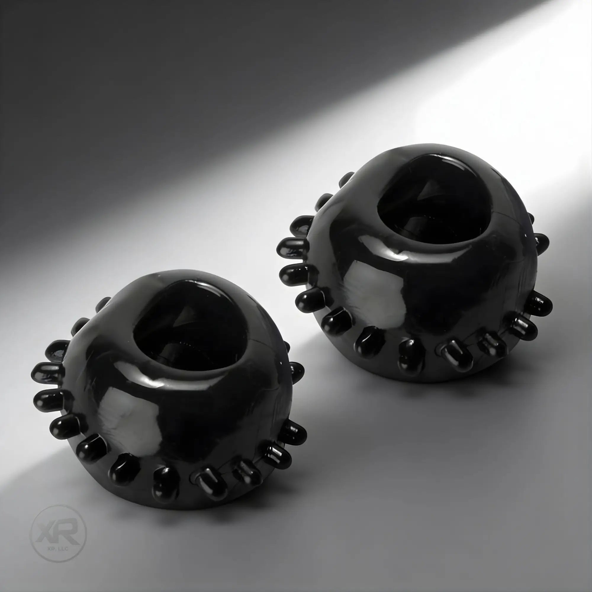 Two shiny black Trinity Vibes gummy cock rings with small textured studs, displayed against a grey background.