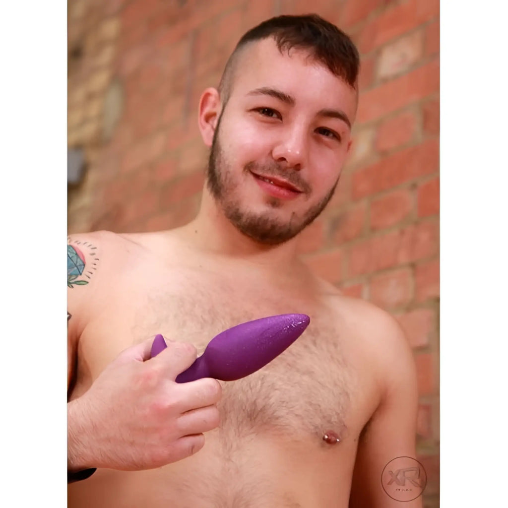 A man holding a purple, tapered Vogue Aria vibrating silicone butt plug against a red brick background.