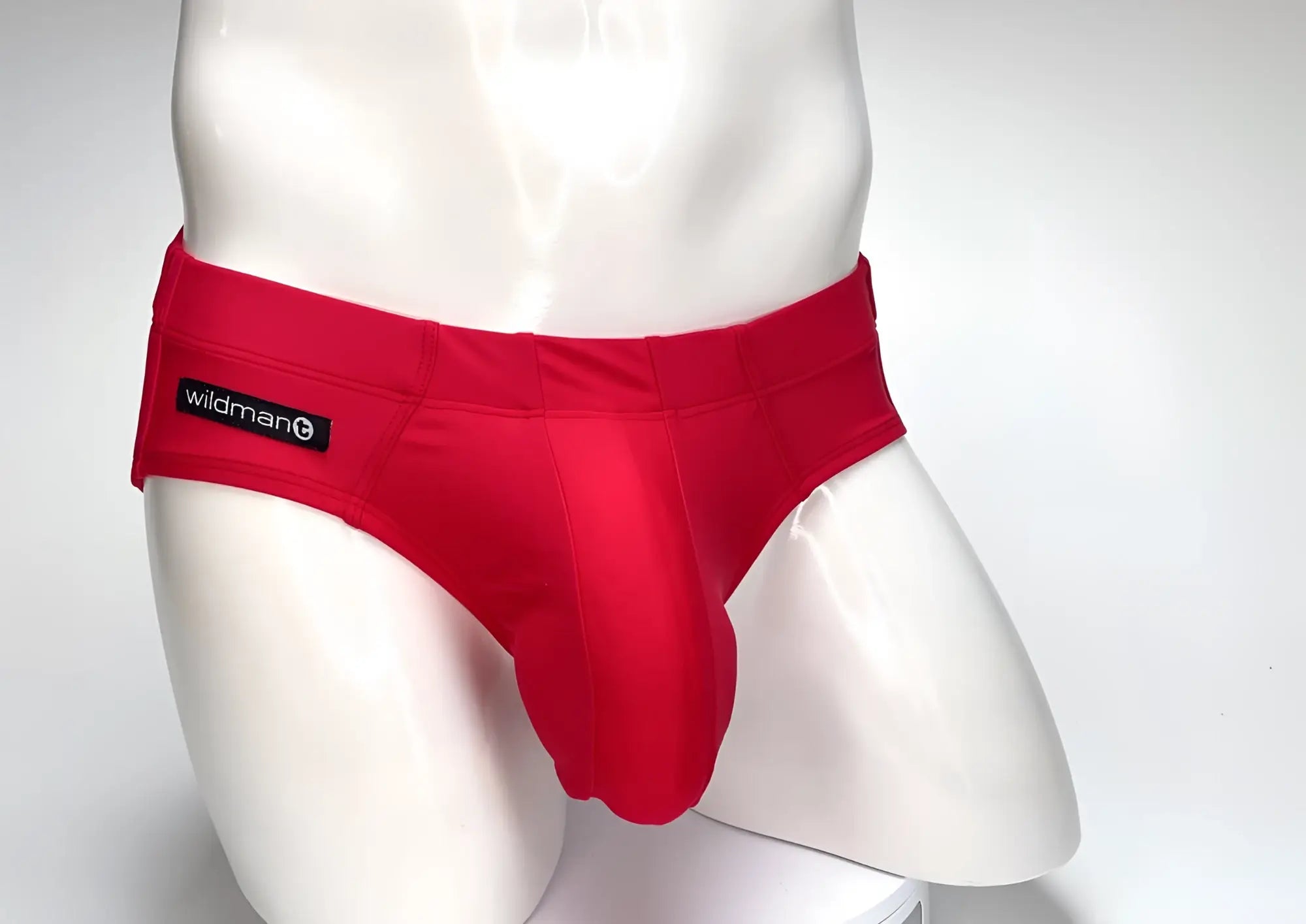 Bright red WildmanT swim brief with a prominent front pouch on a white mannequin torso, featuring a logo patch on the hip.