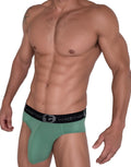 Muscular man wearing sage green WildmanT cotton briefs with a black branded waistband on a white background.