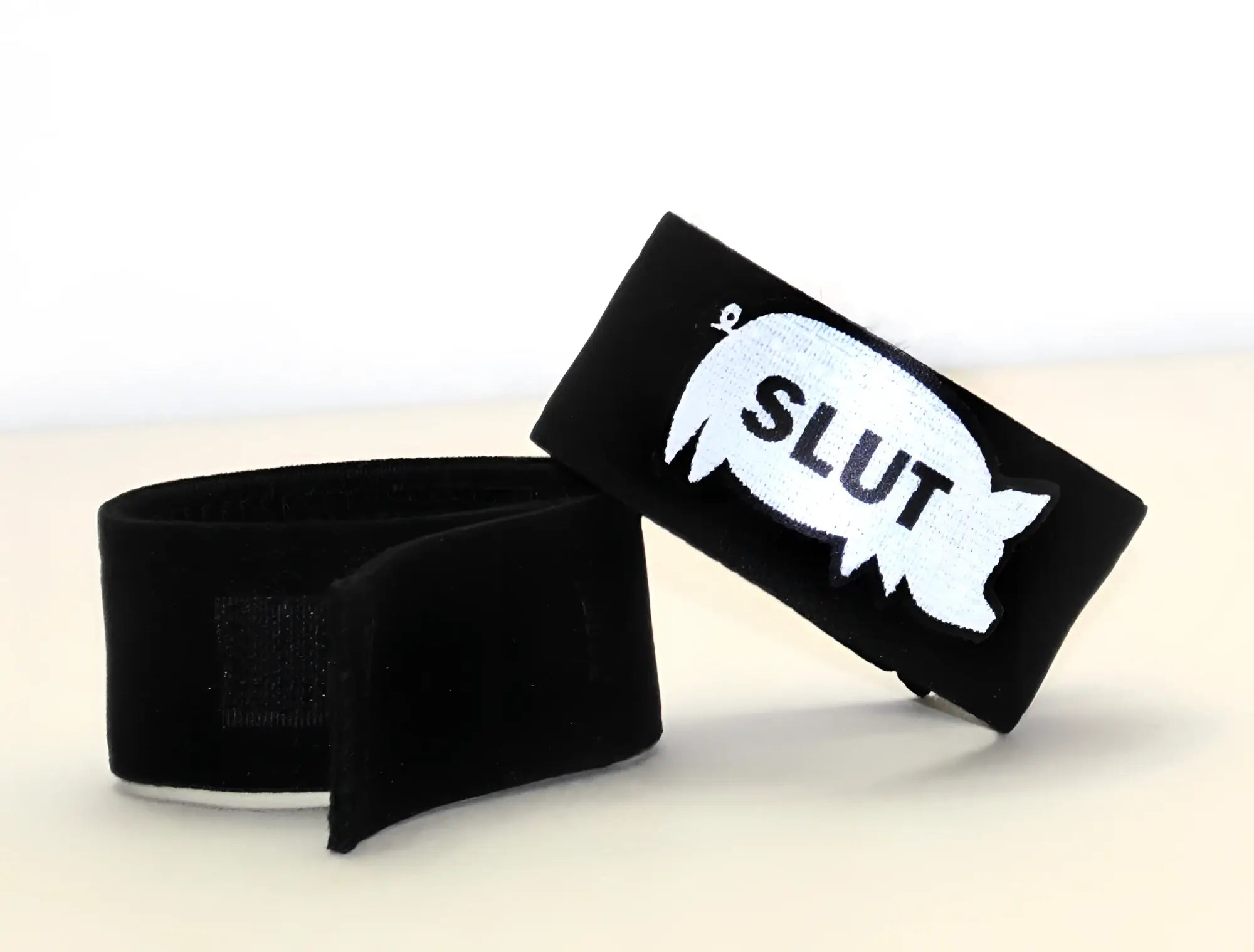 Two black fabric cock rings with hook-and-loop fasteners; one features a white pig silhouette with "SLUT" printed.