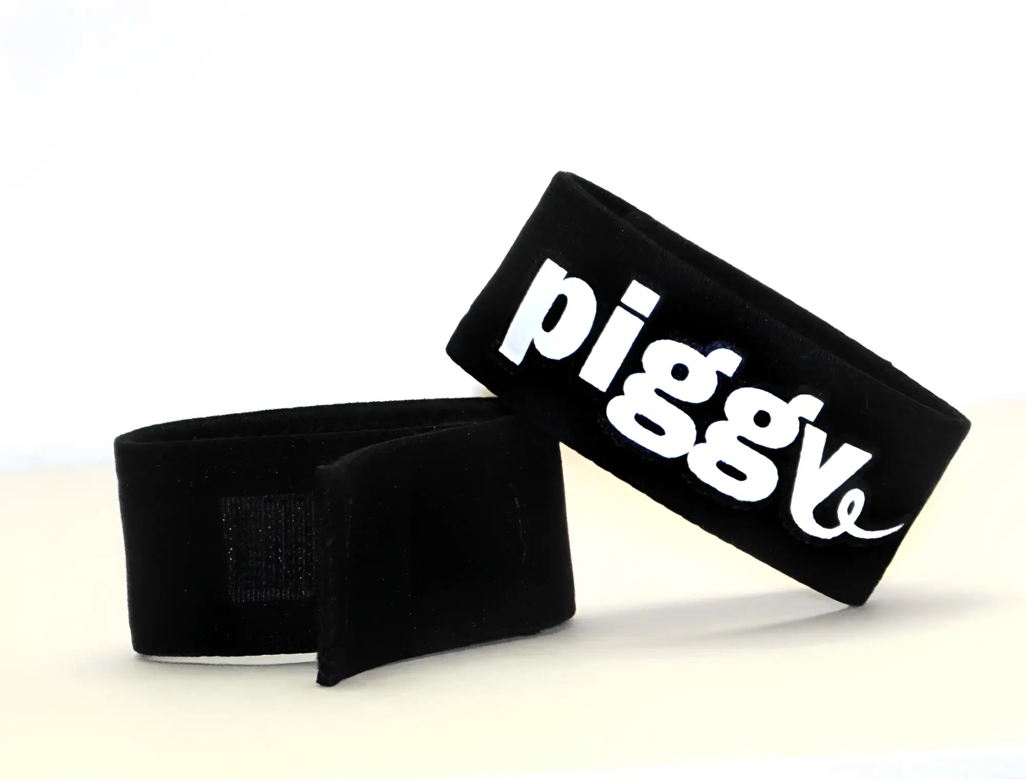 Black adjustable fabric cock ring with a velcro strap and white "piggy" text embroidery on a white background.