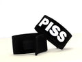 Two black fabric cock rings with velcro closures, featuring the word "PISS" in bold white lettering.
