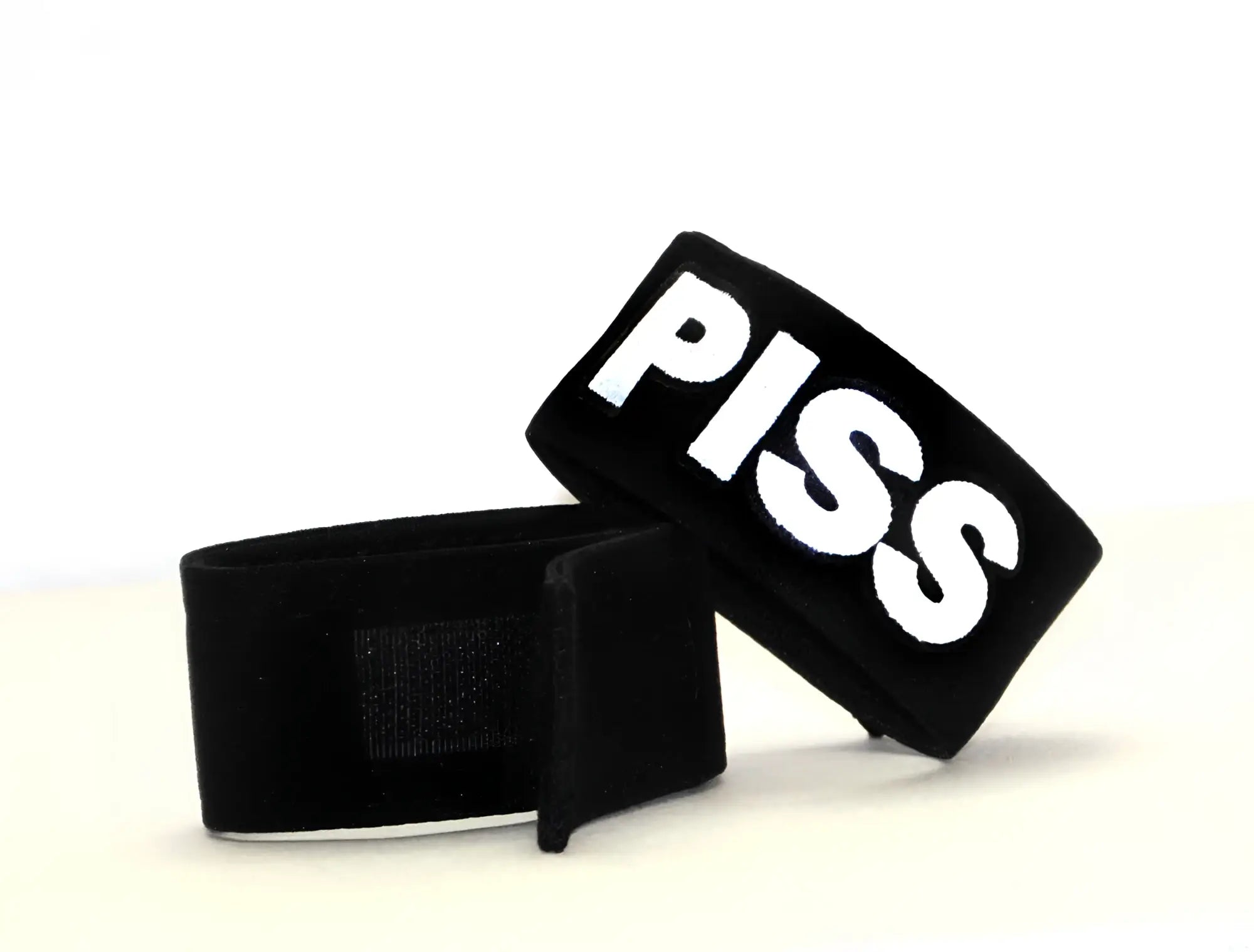 Two black fabric cock rings with velcro closures, featuring the word "PISS" in bold white lettering.